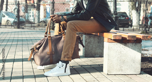 Fashion stylish man with bag sitting on a bench in the city park close up