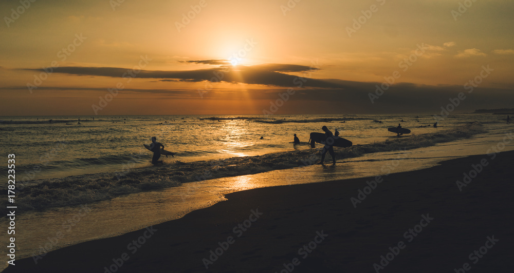 Outstanding ocean view at the nightfall with silhouettes of surfers. Colorful sunset at the tropical sand beach. Group of surfers is spending summer vacation riding waves at the tropical country. Outstanding ocean view at the nightfall with silhouettes of surfers. Colorful sunset at the tropical sand beach. Group of surfers is spending summer vacation riding waves at the tropical country.