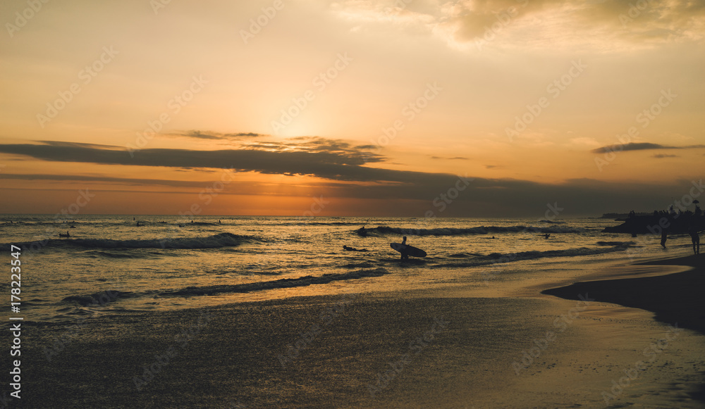 Outstanding sea view at the nightfall with silhouettes of surfers. Colorful dawn at the tropical sand beach. Young surfers are spending summer vacation riding waves at the tropical country.