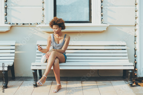Charming young black girl is sending online message via smartphone while sitting on street bench with facade building behind her with copy space zone for text, your advert or other information