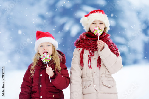 Two adorable little sisters wearing Santa hats having huge striped Christmas lollipops on beautiful winter day © MNStudio
