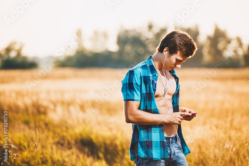 a handsome young man with muscular torso listening to music in white headphones and holding a smartphone in his hand. He is dressed in a shiny blue shirt and jeans shorts, is in a field outside city © Elizaveta