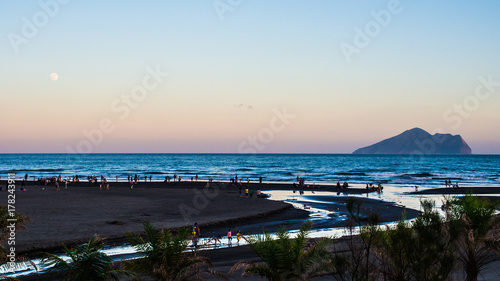 The tourists are enjoyed with black sand and pacific sea of Waiao beach, Taiwan. The sun is setting with vanilla sky, the moon is also showed up on the left side above horizon and the isolated island