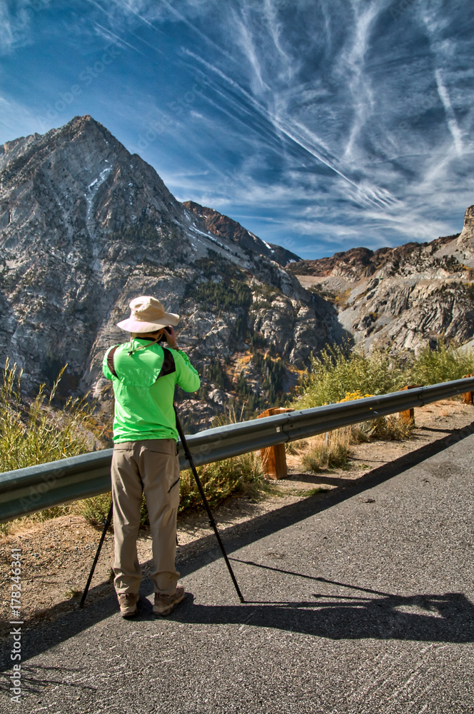 Photographer in a green jacket and white hat takes a picture of the canyon and high granite mountains of Tioga Pass near Yosemite National Park. Blue sky with wispy clouds is in the background. Photographer in a green jacket and white hat takes a picture of the canyon and high granite mountains of Tioga Pass near Yosemite National Park. Blue sky with wispy clouds is in the background.