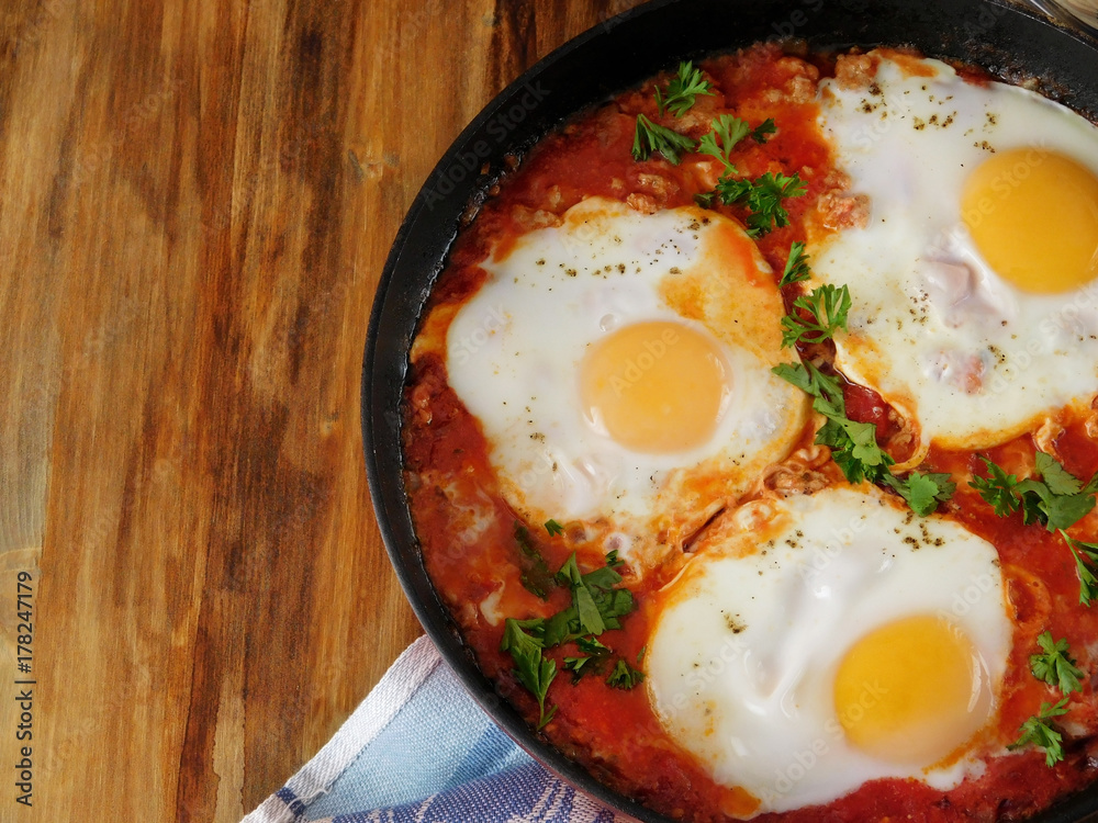 A pan of fried eggs with tomato sauce and parsley on a wooden background. Shakshuka a traditional meal of the Jewish cuisine A pan of fried eggs with tomato sauce and parsley on a wooden background. Shakshuka a traditional meal of the Jewish cuisine