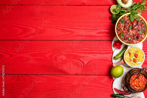 Vegetarian Mexican food concept: refried black and red beans. guacamole, salsa, chili, tortilla chips and fresh ingredients over vintage red rustic wooden background. Top view, flat lay
