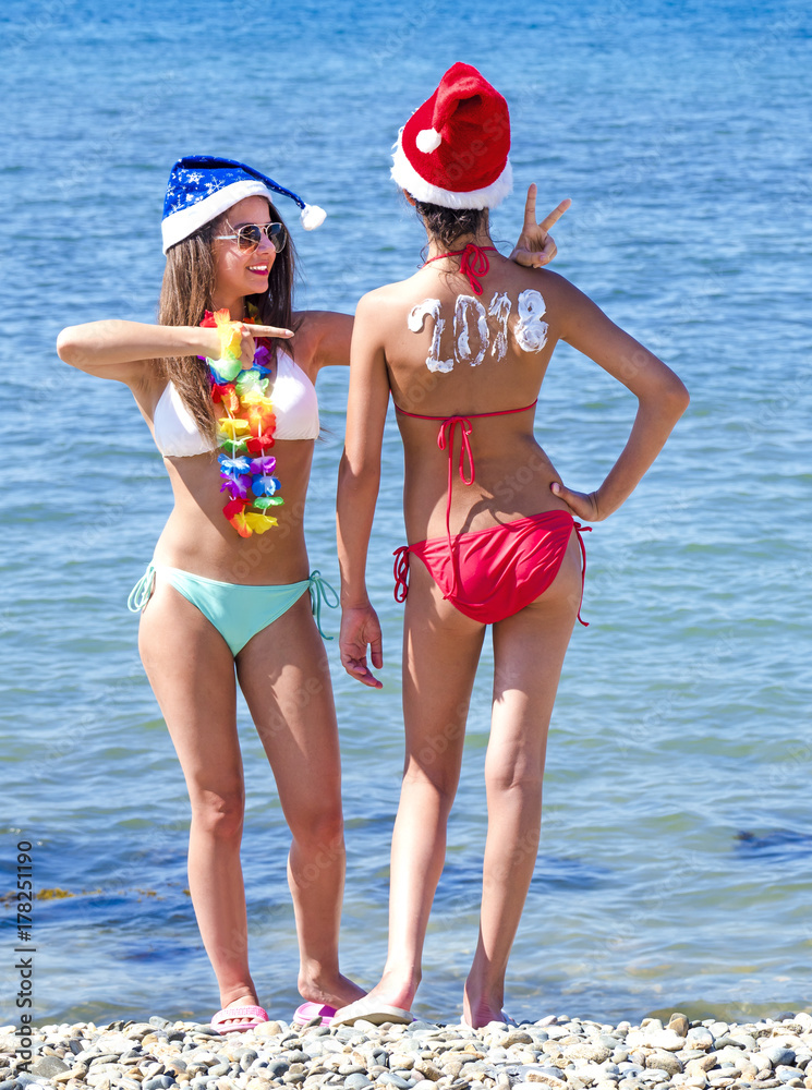 Two girlfriends in a Santa hat during a beach holiday. Girl with the inscription 2018 on the back.  Happy new year