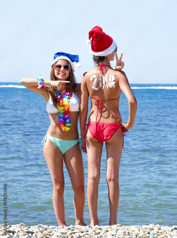 © kravik93 - Two girlfriends in a Santa hat during a beach holiday. Girl with the inscription 2018 on the back.  Happy new year