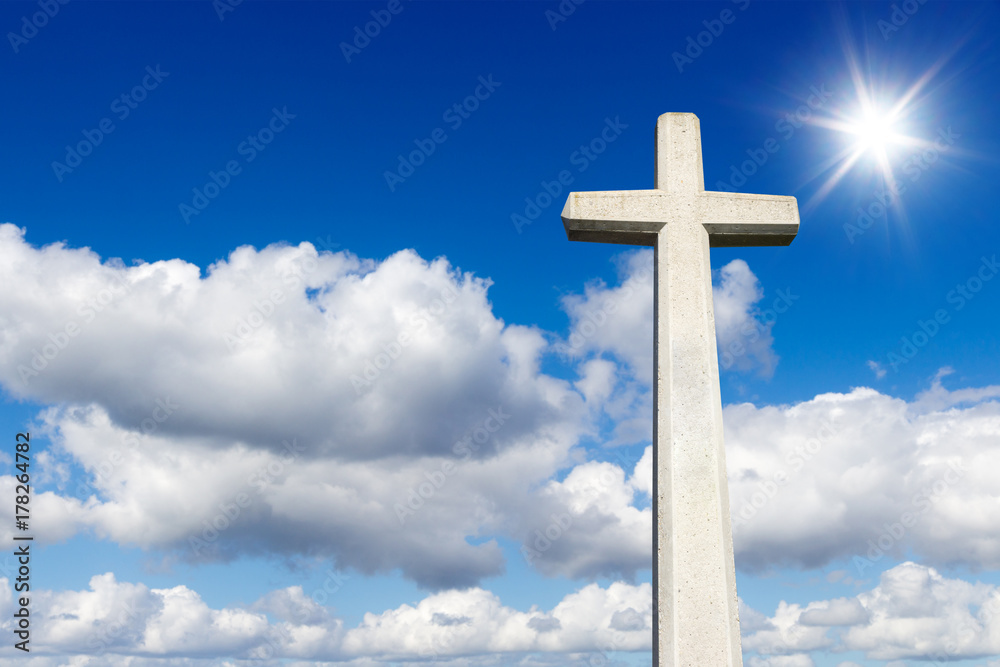 Steinkreuz vor blauem Himmel mit Wolken und der Sonne