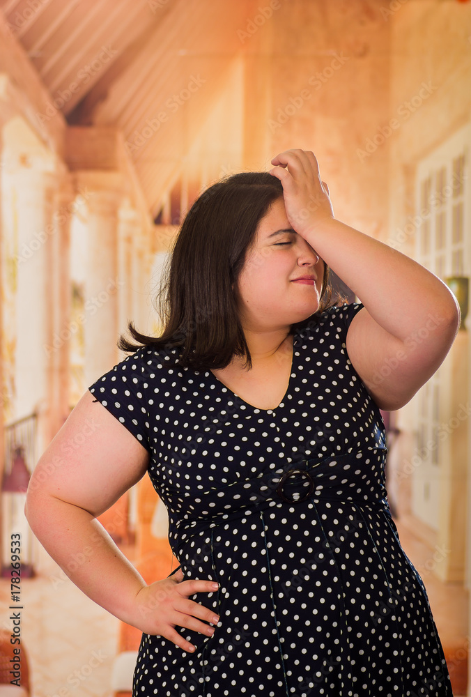 Fototapeta premium Close up of young worried beautiful plus size model in black dres, with one hand in her head and her other in her waist, xxl woman in blurred studio background