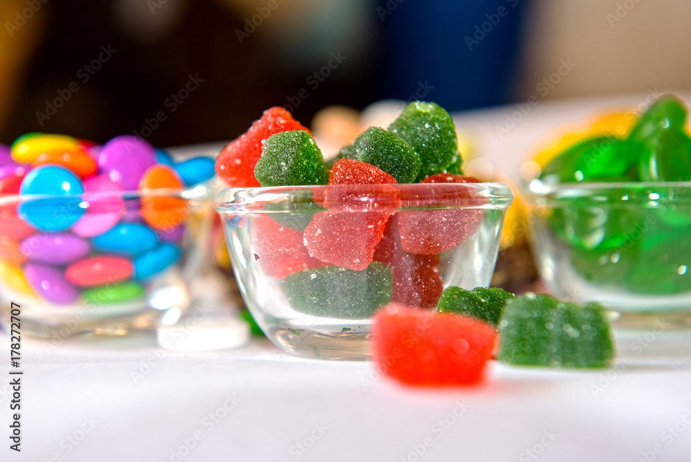 A glass bowl filled with red and green sugar-coated jelly bear candies. Surrounding it are bowls of colorful sweets, including candy-coated chocolates and green hard candies, on a white surface A glass bowl filled with red and green sugar-coated jelly bear candies. Surrounding it are bowls of colorful sweets, including candy-coated chocolates and green hard candies, on a white surface