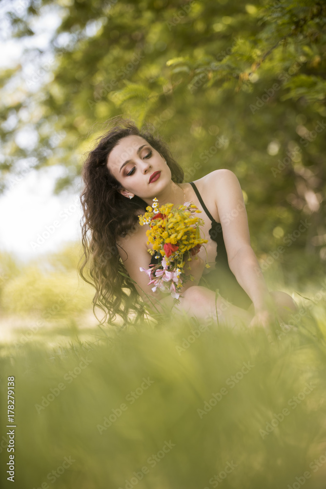 Pretty young lady wear black long dress with deep neckline sitting in the field under the trees holding a bouquet of wild flowers Pretty young lady wear black long dress with deep neckline sitting in the field under the trees holding a bouquet of wild flowers