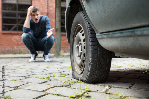 Crouched Man Pointing At Punctured Car Tire