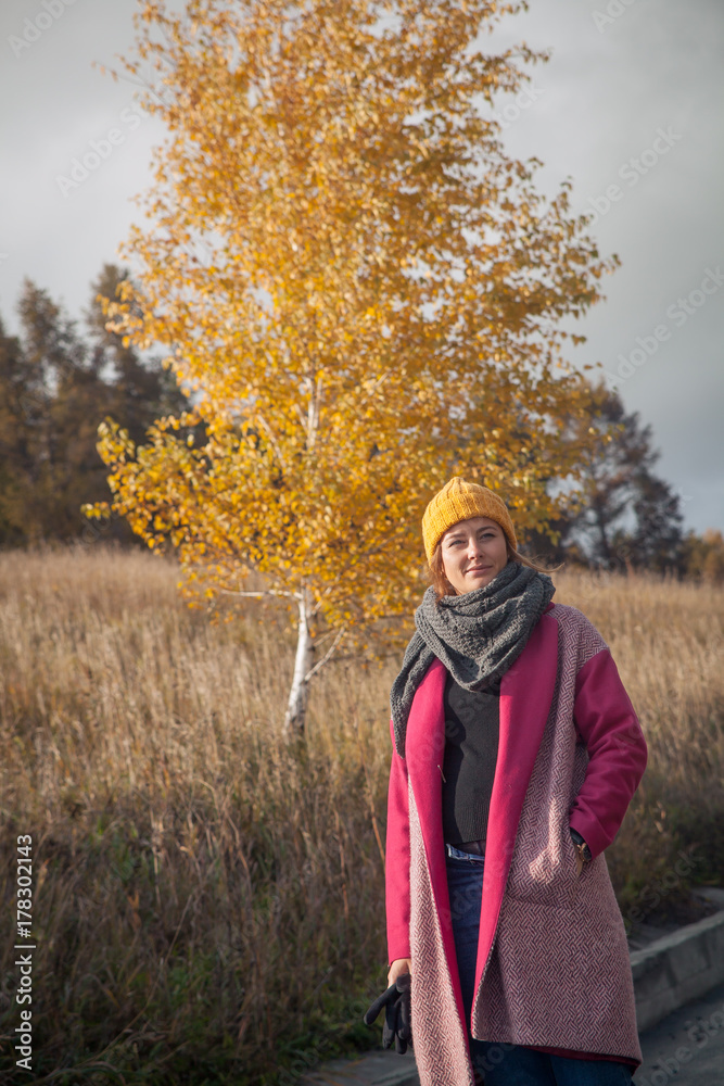 A dark-haired woman in a yellow knitted hat, a knitted gray scarf, a pink coat is standing on the path near the wheat field on an autumn  day, in the background of a birch tree with yellow leaves