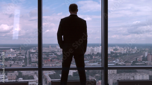Full length back view of successful businessman in suit standing in office with hands on waist, CEO. Businessman from the back in front of a city view on the window photo