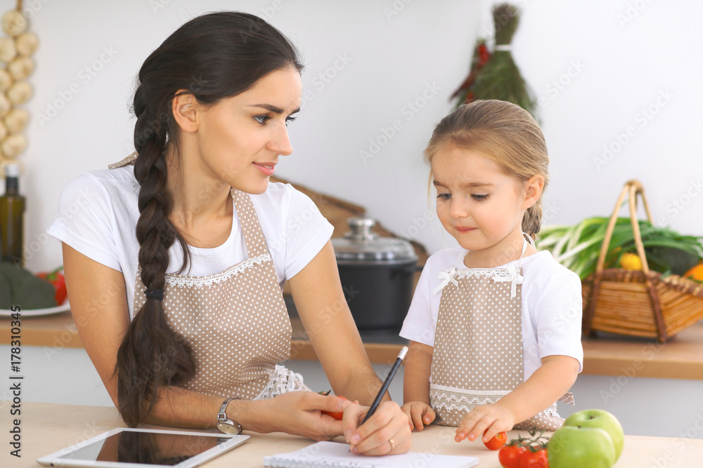 Happy family in the kitchen. Mother and child daughter make menue for cooking tasty breakfest in the kitchen. Little helper is ready for houshold job Happy family in the kitchen. Mother and child daughter make menue for cooking tasty breakfest in the kitchen. Little helper is ready for houshold job