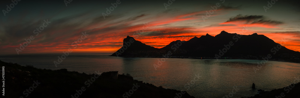 Mountain peak silhouetted against the red sky of sunset with a calm ocean in the foreground.  The Sentinel Peak, Hout Bay, South Africa