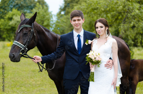 Bride and groom are standing in the park near the horse, wedding walk. White dress, happy couple with an animal. Green background