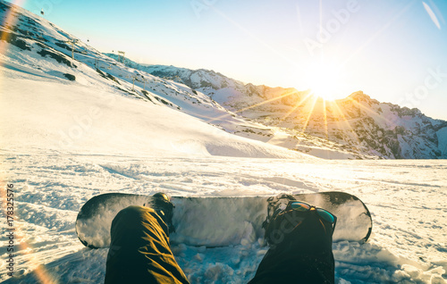 Snowboarder sitting at sunset on relax moment in french alps ski resort - Winter sport concept with adventure guy on top of mountain ready to ride down - Legs view point with teal and orange filter