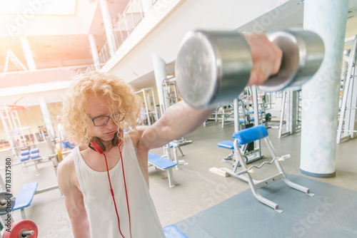 Weak curly blonde man holding a dumbbell with a strained face in the sunlight
