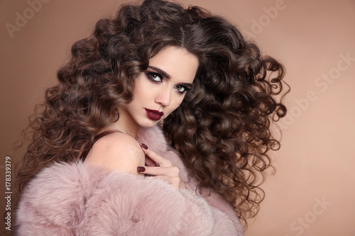 Hairstyle. Beauty hair. Fashion brunette girl with long curly hair, beauty makeup. Glamour portrait of beautiful woman with marsala matte lips in pink fur coat isolated on beige background.