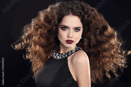Hairstyle. Fashion brunette girl with Long curly hair, beauty makeup. Glamour portrait of beautiful woman with marsala matte lips, blowing hairstyle isolated on black background.