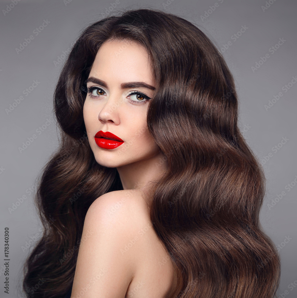Fototapeta premium Beauty hair. Brunette girl portrait with red lipstick and long shiny wavy hair. Beautiful model with healthy hairstyle isolated on studio dark background.