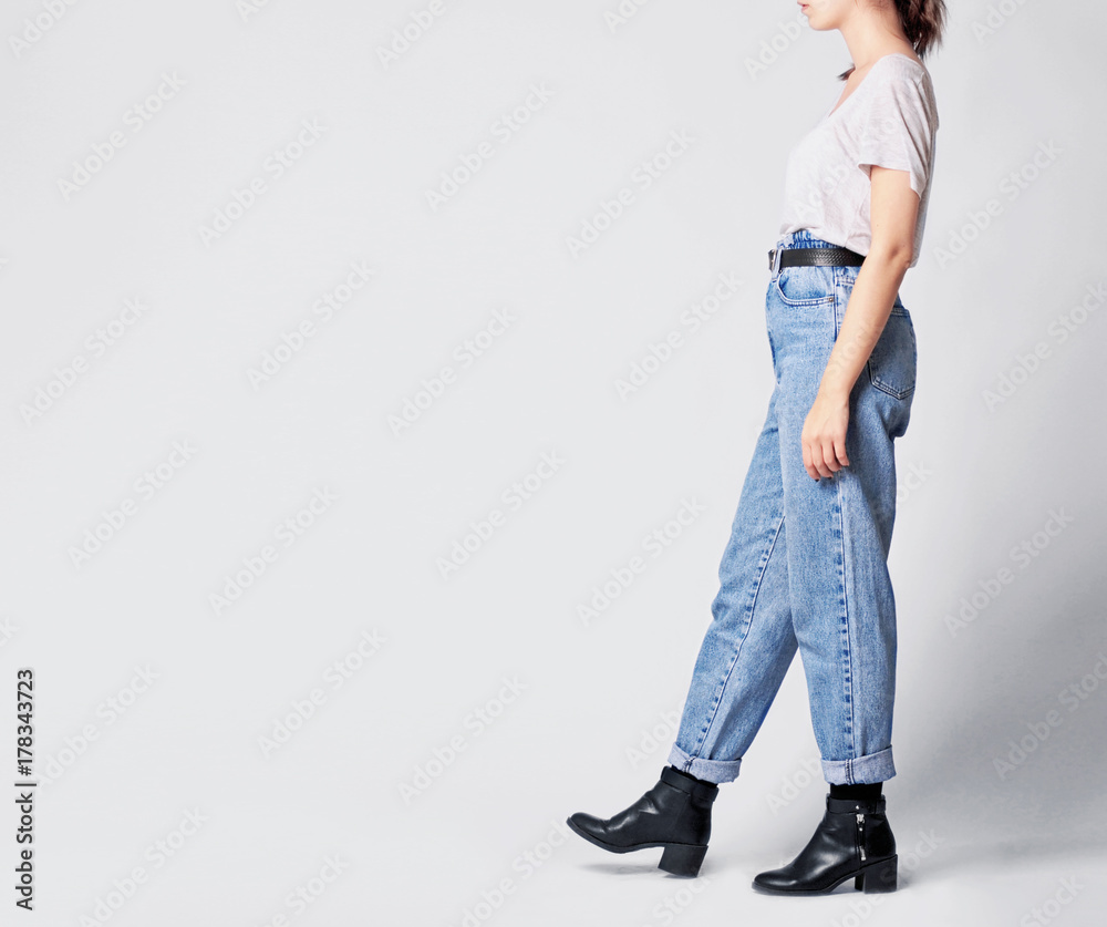 Woman wearing casual outfit with white t-shirt, blue vintage high-waisted mom jeans with black faux leather ankle boots and black belt isolated on grey background. Copy space Stock Photo Adobe Stock