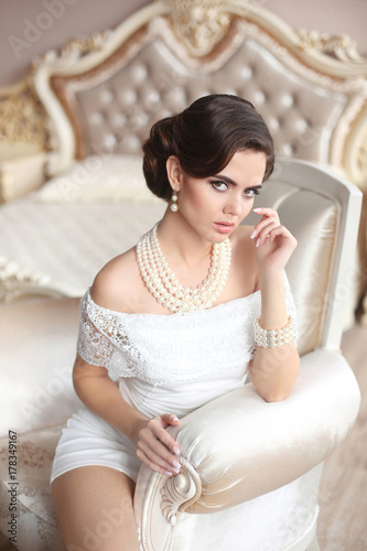 Retro woman portrait. Elegant brunette lady with fashion pearls jewelry set, wavy hairstyle and makeup. Gorgeous model posing on luxury armchair in modern interior.