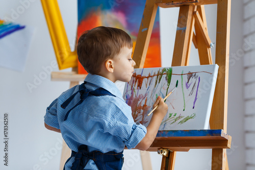Fototapeta Naklejka Na Ścianę i Meble -  Cute, serious and focused, three years old boy in blue shirt and jeans apron painting on canvas standing on the easel. Concept of early childhood education, talent, happy family or parenting