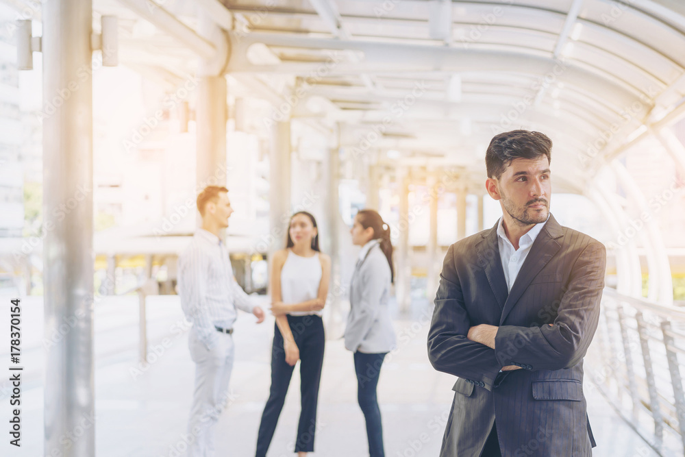 Portrait of handsome business man standing with arms crossed in outdoors. Smart caucasian young manager with his colleagues in background. Portrait of handsome business man standing with arms crossed in outdoors. Smart caucasian young manager with his colleagues in background.