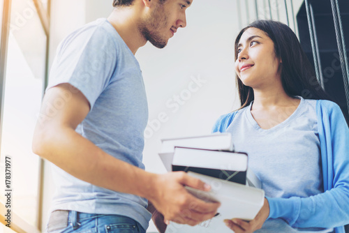 Two young students holding books and both of them catch eye contact. Happy young couple smiling and reading book in library.