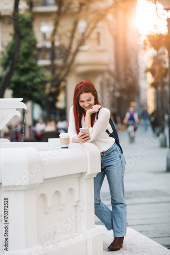 Young woman standing at the street next to drinking fountain using mobile phone and drinking coffee to go 