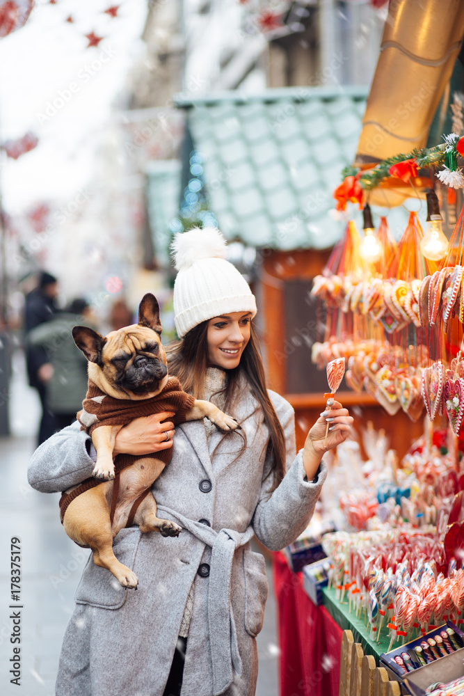 Beautiful brunette woman standing with her adorable French bulldog next to candies selling on street.