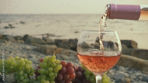 Slow mo rose wine from a bottle is filled in a glass on a background of a sea stony coast with bunches of grapes