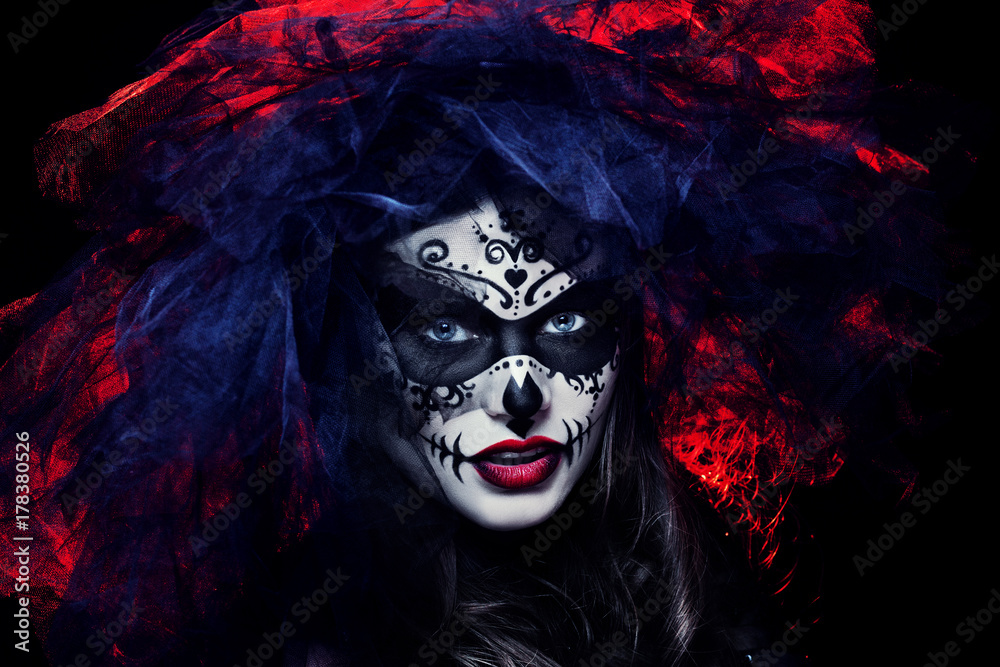 Close up studio portrait of beautiful woman with Halloween sugar skull makeup in red and black colors, wearing bridal veil. Model looking at camera. Dark, dead bride. 
