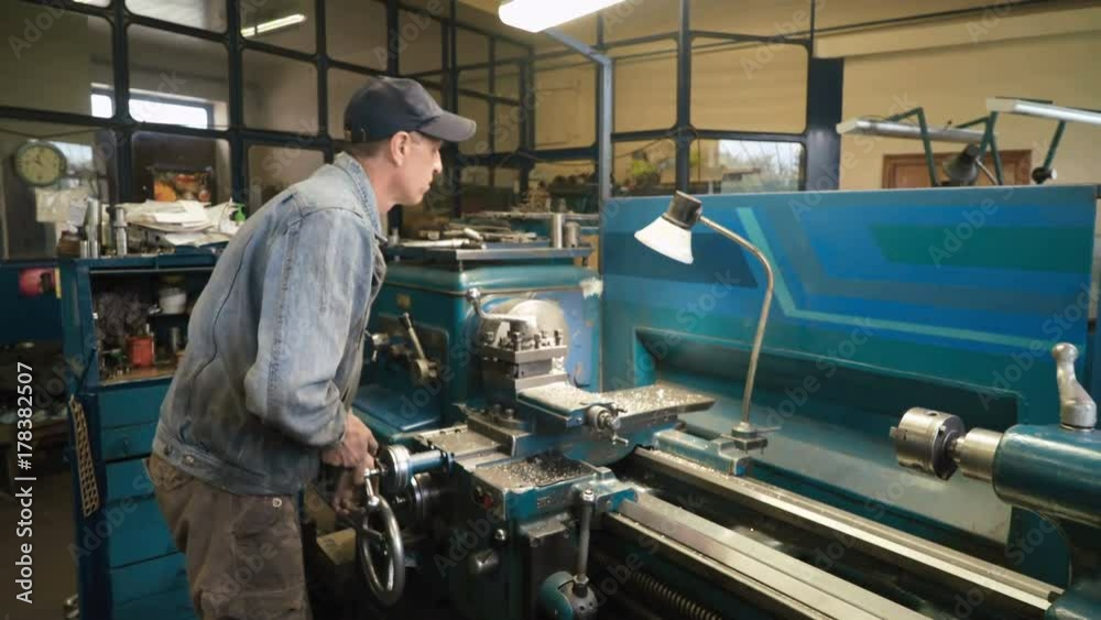 Mature caucasian man working in shabby car service, making aluminum bushing on old lathe. Wearing blue jeans jacket and cap. Shot with Sony a7s and Atomos Ninja Flame indoors, dolly slide Mature caucasian man working in shabby car service, making aluminum bushing on old lathe. Wearing blue jeans jacket and cap. Shot with Sony a7s and Atomos Ninja Flame indoors, dolly slide