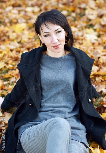 girl in black coat sitting on a yellow leaves in autumn city park, fall season