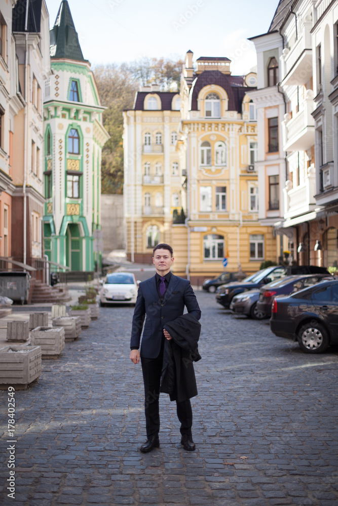 Portrait of a serious fashionable handsome man in a blue suit and tie is holding a coat in the streets of the city, vertically.