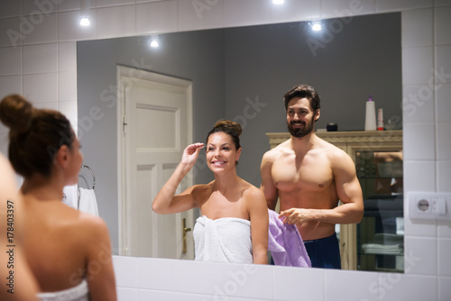 Konstfotografi Young cute girl plucking eyebrows depilating with tweezers in front of a strong muscular shirtless man whole holding towel in the bathroom