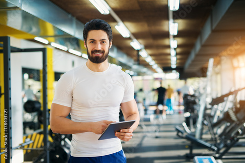 Muscular handsome bearded trainer looking at the camera while standing with a tablet in the gym.