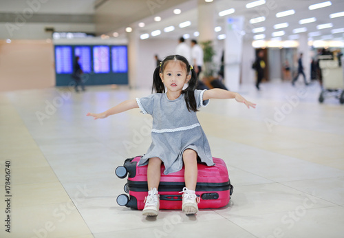 Adorable little asian girl at airport sitting on suitcase with open arm.