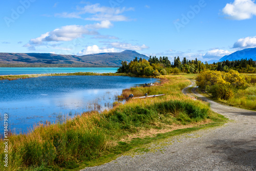 Gravel road between a marsh and lake in the Alaskan landscape, with mountains and a cloudy sky in the background 