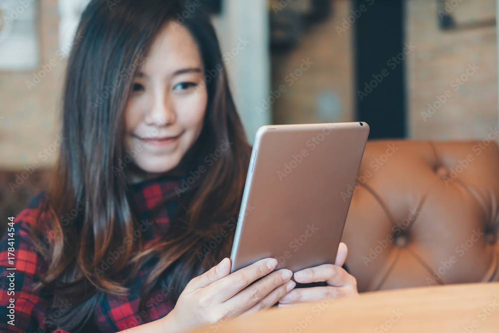 Closeup image of a smiley beautiful Asian woman holding ,using and looking at tablet pc sitting in modern cafe Closeup image of a smiley beautiful Asian woman holding ,using and looking at tablet pc sitting in modern cafe