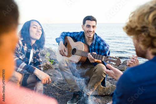 Happy guy with guitar and his friends spending evening around campfire by seaside