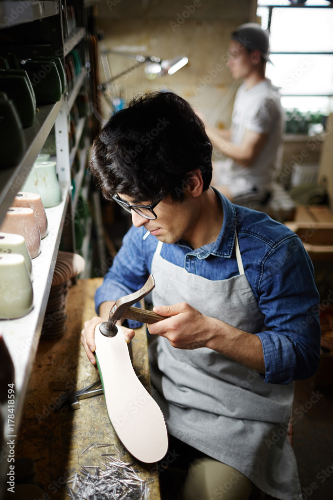 Young man in apron sitting by his workplace and hammering nails into sole of footwear workpiece