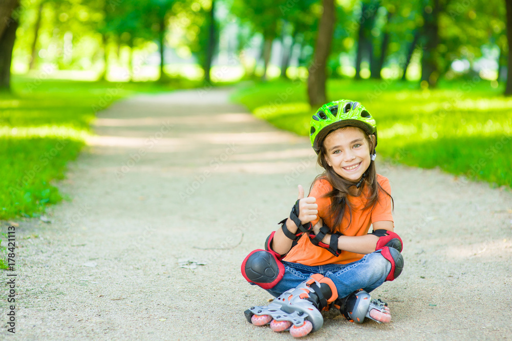 Happy girl in a protective helmet and protective pads for roller skating sitting on the road and showing thumbs up. Space for text Happy girl in a protective helmet and protective pads for roller skating sitting on the road and showing thumbs up. Space for text