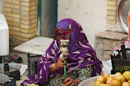 Iranian woman in traditional colorful mask sitting on local bazaar and smoking water pipe. photo