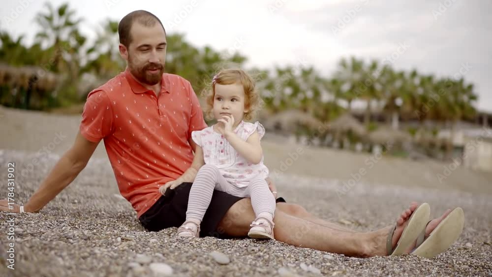 cute little girl sitting at her beloved dad on his feet and waving at the camera with your hand. a bearded man resting his daughter on the sea sitting on the beach at sunset cute little girl sitting at her beloved dad on his feet and waving at the camera with your hand. a bearded man resting his daughter on the sea sitting on the beach at sunset