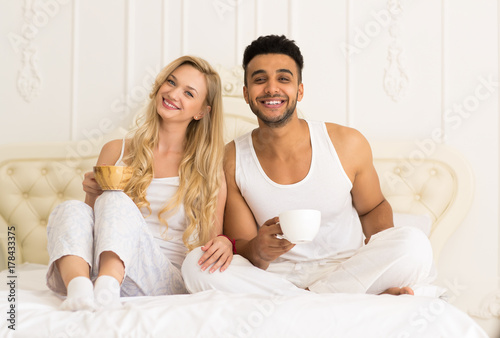 Young Couple Drink Coffee Orange Juice Sitting In Bed, Happy Smile Young Hispanic Man And Woman Lovers Hold Cups Glasses Bedroom © mast3r